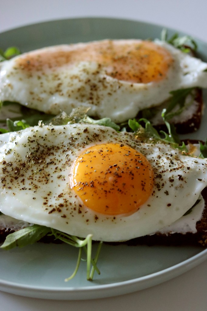 Two sunny side up fried eggs on dark toast with arugula, cream cheese, and black pepper